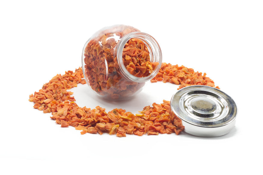 A jar of carrot flakes next to a small, open bowl of the same product, with some carrot flakes scattered around.
